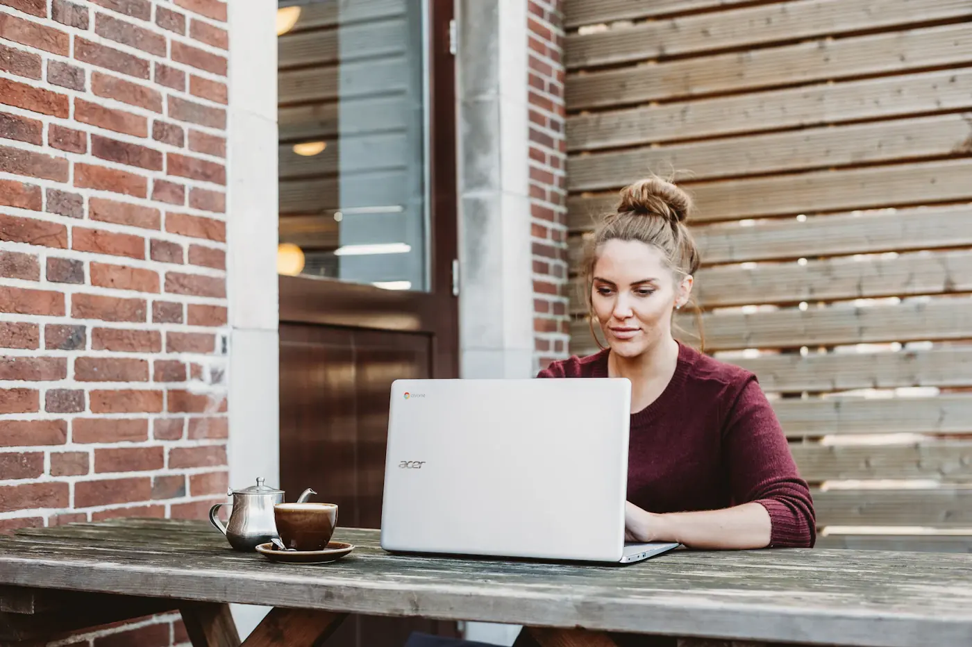 A woman working on a laptop at an outdoor table against a brick wall