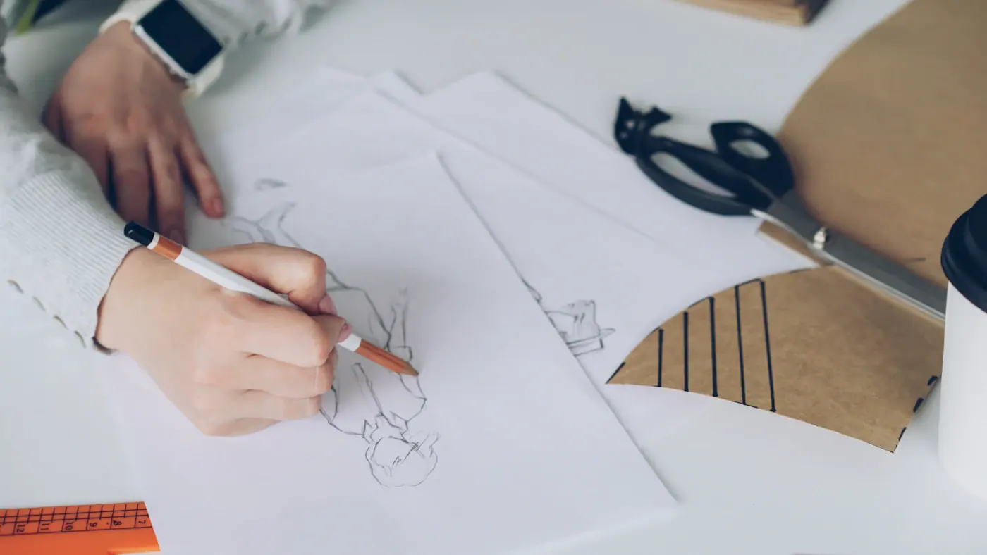A hand sketching a design on paper next to a pair of scissors